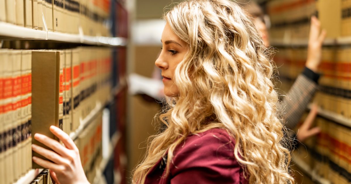 Blonde student with curly hair and maroon shirt picking out a book in the library.