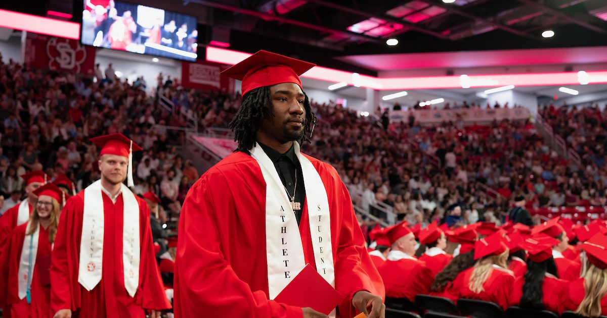 A student athlete wearing a red cap and gown walking at graduation ceremony.
