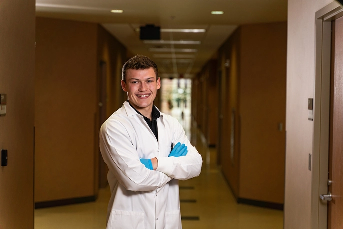 A medical student dressed in a white lab coat and blue gloves standing in the hallway of a hospital.