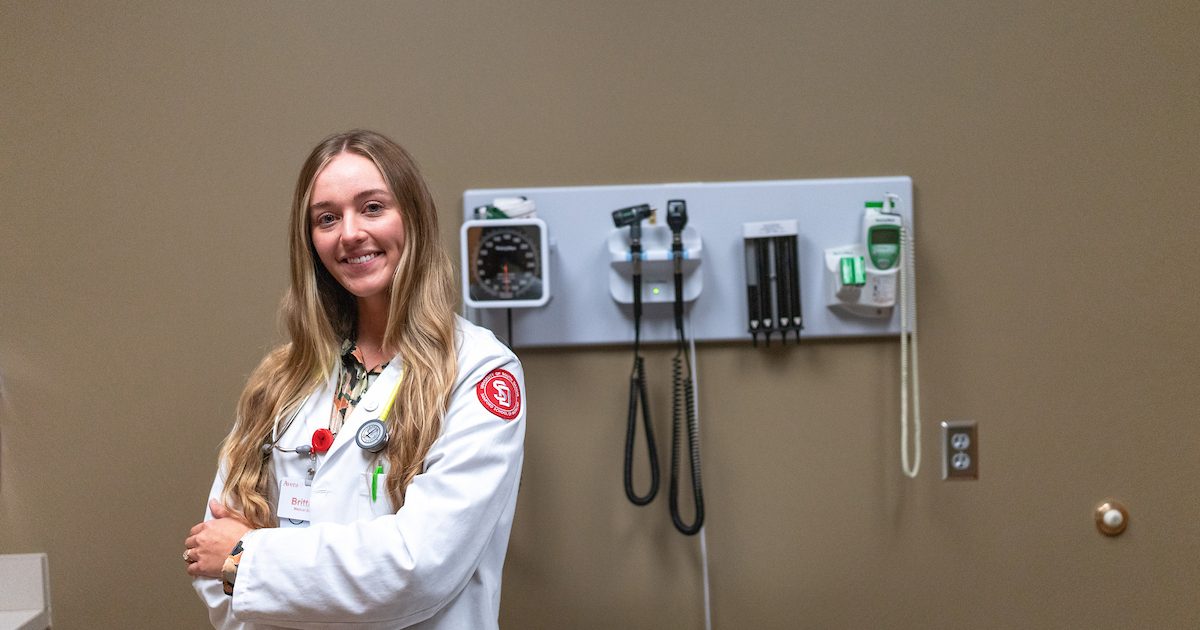 Medical student in white coat posing in front of medical tools in a doctors office.