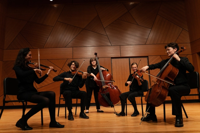Five string orchestra members performing on stage wearing all black.